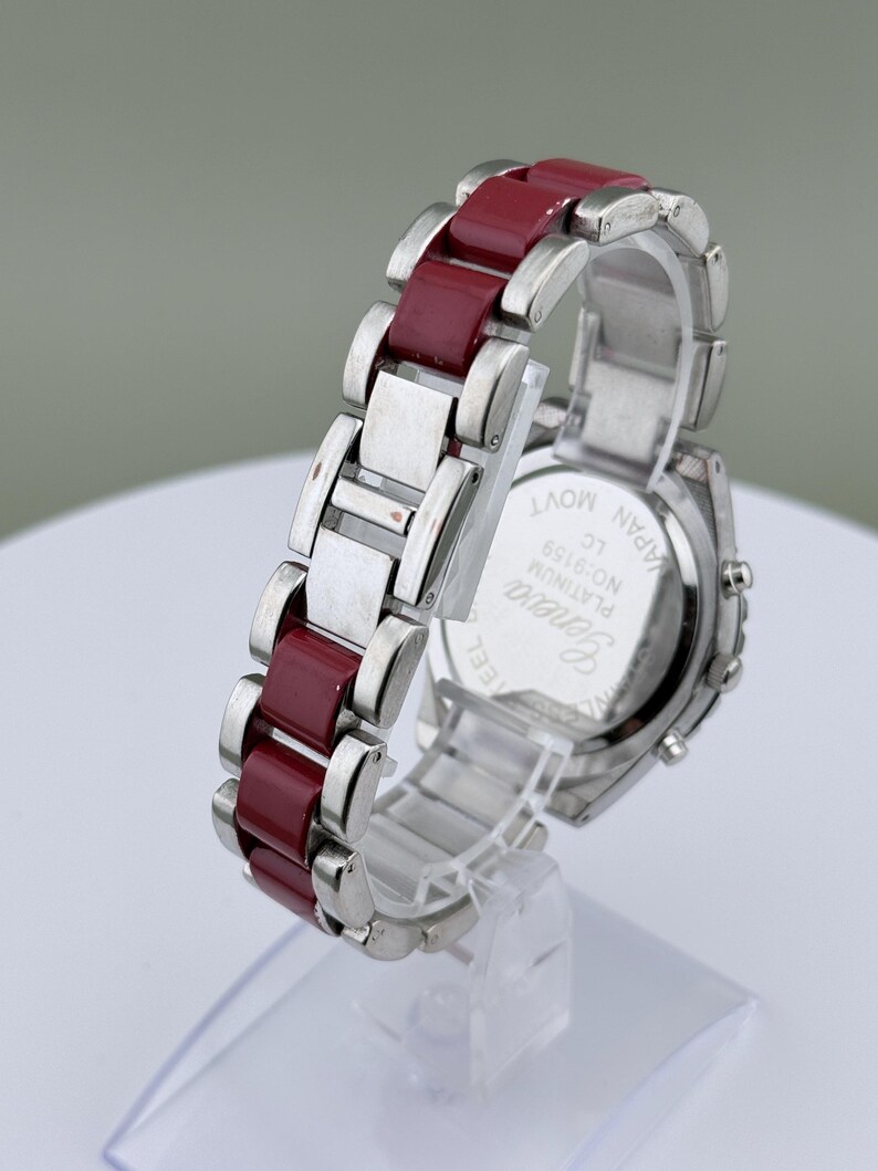 May include: A silver-toned wristwatch with a burgundy band. The watch features a rectangular face and a stainless steel band with burgundy links. The watch is displayed on a clear stand.