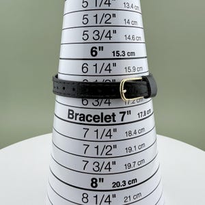K&ouml;nnte beinhalten: Ein schwarzes Lederarmband mit goldfarbener Schnalle wird auf einem wei&szlig;en Armband-Messkegel gezeigt. Der Kegel zeigt Messungen in Zoll und Zentimetern. Das Armband passt bei 17,8 cm.