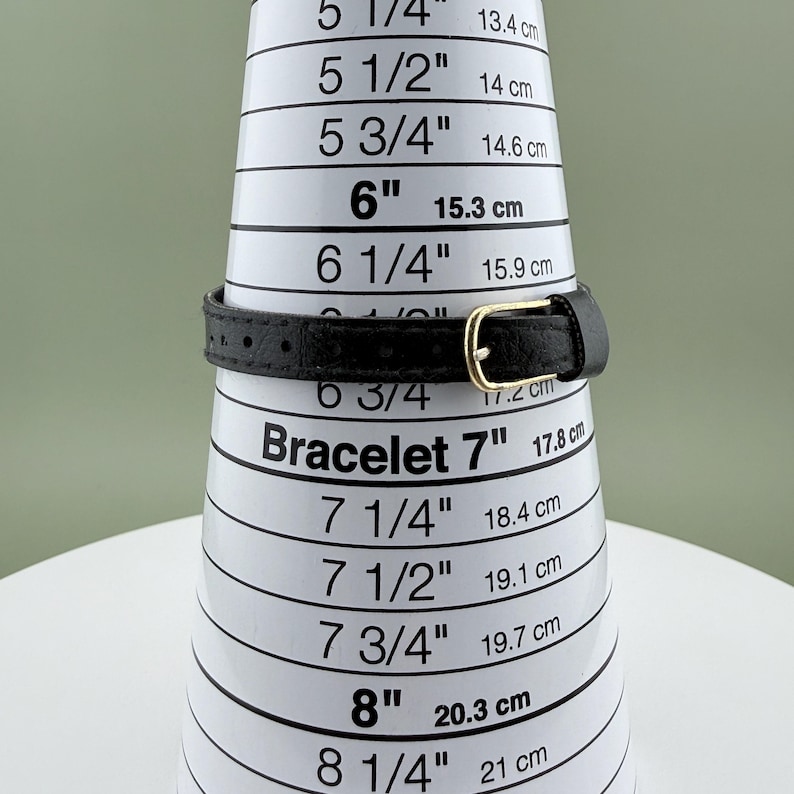 K&ouml;nnte beinhalten: Ein schwarzes Lederarmband mit goldfarbener Schnalle wird auf einem wei&szlig;en Armband-Messkegel gezeigt. Der Kegel zeigt Messungen in Zoll und Zentimetern. Das Armband passt bei 17,8 cm.