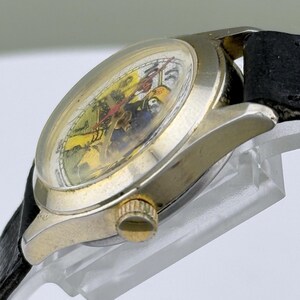 K&ouml;nnte beinhalten: Eine Vintage-Armbanduhr mit goldfarbenem Geh&auml;use und schwarzem Lederarmband. Das Zifferblatt der Uhr hat ein farbenfrohes, detailliertes Design mit roten, gelben und blauen Elementen. Die Uhr ist abgewinkelt und zeigt die Seite und das Zifferblatt.