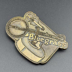 May include: A bronze-colored metal pin featuring a banjo, guitar, and violin. The word "Bluegrass" is embossed on the pin. The pin has a detailed design and is likely a collectible item or a decorative accessory.