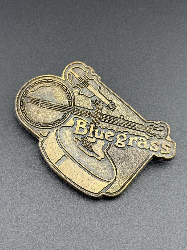 May include: A bronze-colored metal pin featuring a banjo, guitar, and violin. The word "Bluegrass" is embossed on the pin. The pin has a detailed design and is likely a collectible item or a decorative accessory.