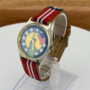 May include: A wristwatch with a patriotic theme. The watch face features the Statue of Liberty and the American flag against a blue background with stars. The watch has a gold-colored case and a red, white, and blue striped band with a brown leather backing.