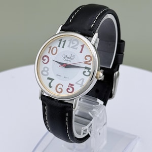 May include: A silver-toned wristwatch with a white face and a black leather band. The watch face features colorful numerals and the brand name "Swednic Quartz". The watch is displayed on a clear stand.