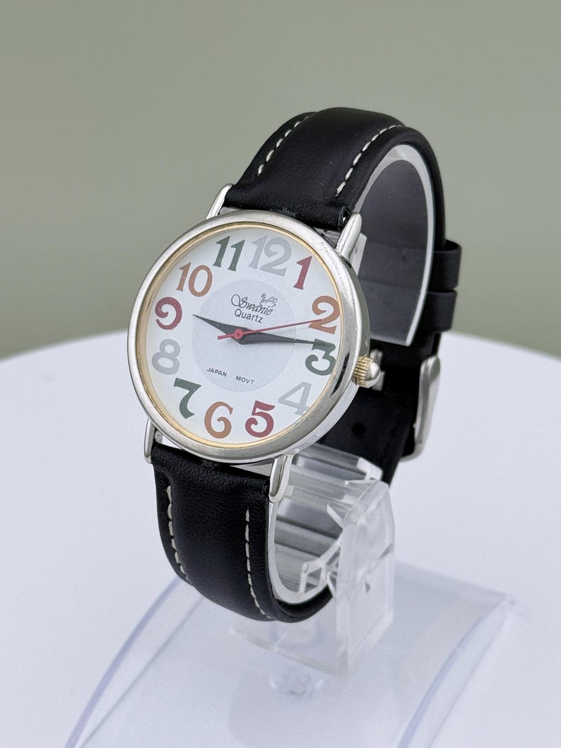 May include: A silver-toned wristwatch with a white face and a black leather band. The watch face features colorful numerals and the brand name "Swednic Quartz". The watch is displayed on a clear stand.
