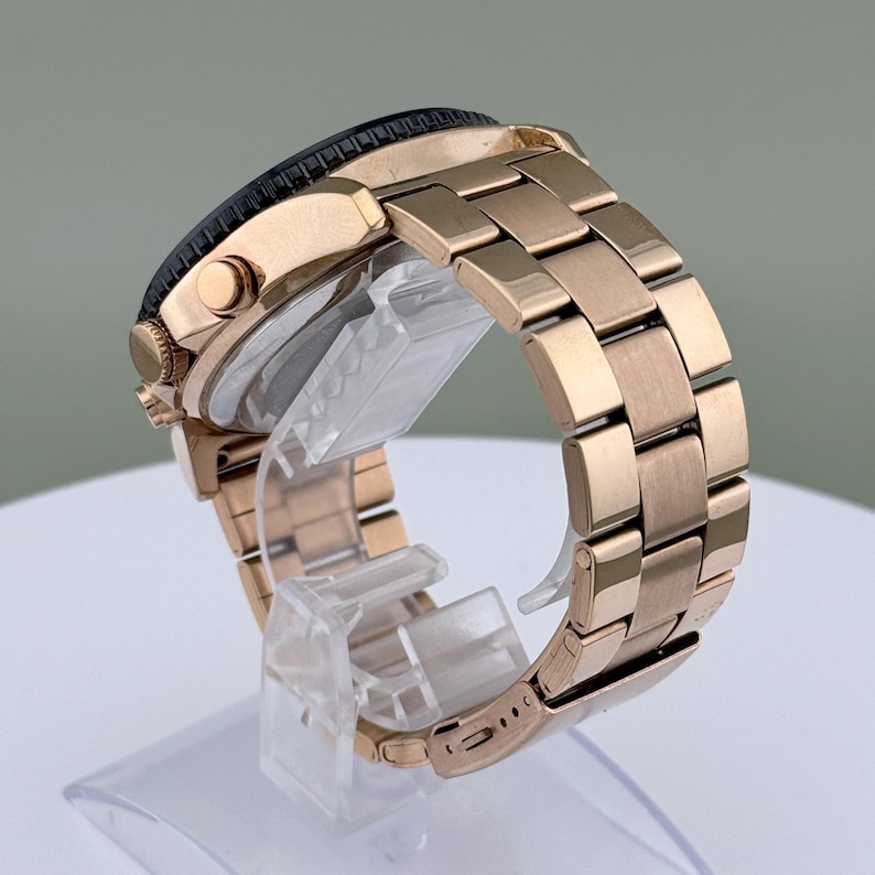 K&ouml;nnte beinhalten: Eine ros&eacute;goldfarbene Armbanduhr mit schwarzer L&uuml;nette und passendem Metallarmband. Die Uhr wird auf einem transparenten St&auml;nder pr&auml;sentiert, der ihr Seitenprofil und die detaillierten Glieder des Armbands hervorhebt.