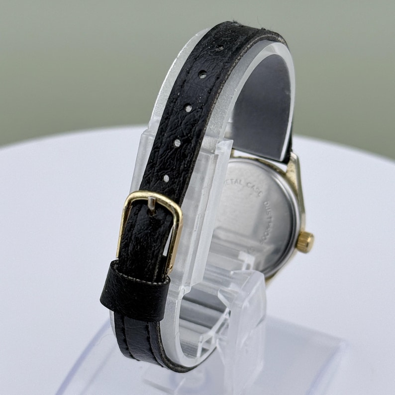 K&ouml;nnte beinhalten: Eine Vintage-Armbanduhr mit einem schwarzen Lederarmband und einem goldfarbenen Geh&auml;use. Die Uhr hat ein klassisches Design mit einem Schnallenverschluss. Die Uhr wird auf einem transparenten St&auml;nder pr&auml;sentiert, der die R&uuml;ckseite der Uhr zeigt.