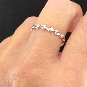 May include: A silver ring with a repeating wave-like design. The ring is worn on a finger, showcasing its delicate, flowing pattern. The ring's design is a series of connected, stylized waves.