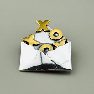 May include: A silver-toned metal pin shaped like an open envelope with gold-colored letters "XO" inside. The pin has a textured surface and a simple, elegant design. The letters are in a bold, sans-serif font.
