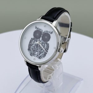 May include: A silver-toned wristwatch with a black leather band. The watch face is white with a black and white owl illustration. The watch has a silver bezel and small crystal accents. The watch is displayed on a clear stand.