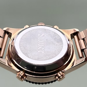 K&ouml;nnte beinhalten: Nahaufnahme einer ros&eacute;goldenen Armbanduhr. Der Uhrenboden ist sichtbar und zeigt den Markennamen "Stauer" sowie "Stainless Steel", "Water Resistant 30M" und die Seriennummer 46954. Das Armband und das Geh&auml;use sind ebenfalls ros&eacute;gold.