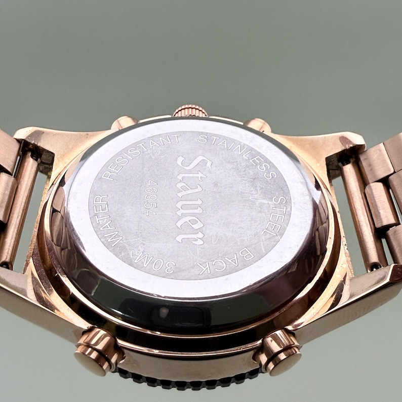 K&ouml;nnte beinhalten: Nahaufnahme einer ros&eacute;goldenen Armbanduhr. Der Uhrenboden ist sichtbar und zeigt den Markennamen "Stauer" sowie "Stainless Steel", "Water Resistant 30M" und die Seriennummer 46954. Das Armband und das Geh&auml;use sind ebenfalls ros&eacute;gold.