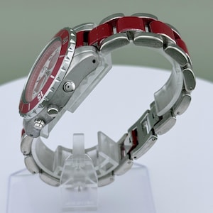 May include: A stainless steel wristwatch with a red bezel and red links on the band. The watch face is red with white markings. The watch is displayed on a clear plastic stand.