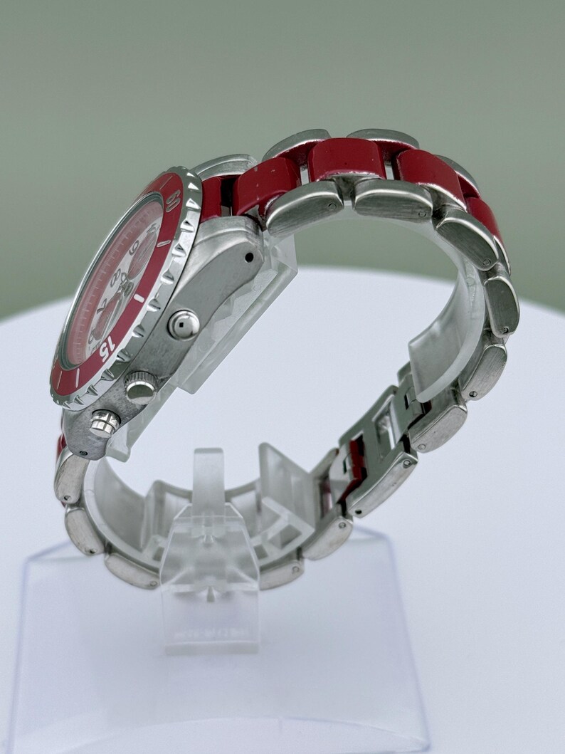 May include: A stainless steel wristwatch with a red bezel and red links on the band. The watch face is red with white markings. The watch is displayed on a clear plastic stand.
