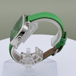 May include: A green watch with a square face and a silver bezel. The watch has a green leather strap. The watch is displayed on a clear stand. The watch face and strap are a matching shade of green.