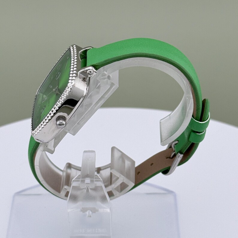 May include: A green watch with a square face and a silver bezel. The watch has a green leather strap. The watch is displayed on a clear stand. The watch face and strap are a matching shade of green.