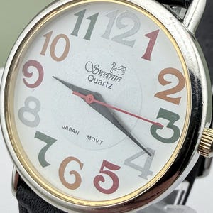 May include: A Swanie Quartz wristwatch with a white face and a black leather band. The watch face features multi-colored numerals and hands, with the words "Swanie Quartz" and "Japan Movt" visible.