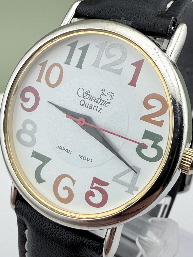 May include: A Swanie Quartz wristwatch with a white face and a black leather band. The watch face features multi-colored numerals and hands, with the words "Swanie Quartz" and "Japan Movt" visible.