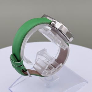 May include: A wristwatch with a silver bezel and a green leather strap. The watch is displayed on a clear plastic stand. The strap is a bright green colour and appears to be leather. The watch face is not visible.
