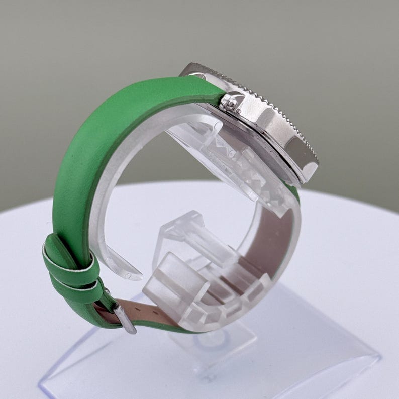 May include: A wristwatch with a silver bezel and a green leather strap. The watch is displayed on a clear plastic stand. The strap is a bright green colour and appears to be leather. The watch face is not visible.