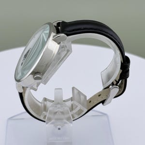 May include: A silver-toned wristwatch with a white face and black hands. The watch has a black leather band and a silver case. The watch is displayed on a clear plastic stand.