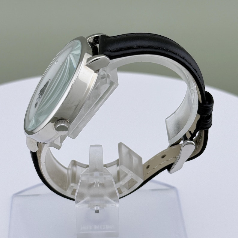 May include: A silver-toned wristwatch with a white face and black hands. The watch has a black leather band and a silver case. The watch is displayed on a clear plastic stand.