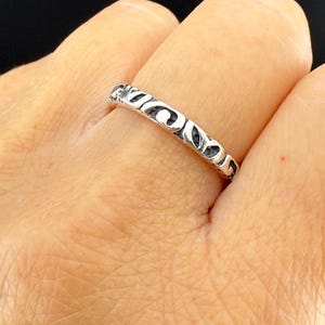 May include: A silver ring with an openwork design featuring a repeating pattern of curved shapes. The ring is worn on a finger, showcasing the intricate details of the metalwork. The ring's design is visible in a close-up shot.