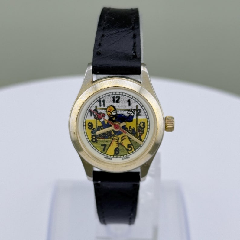 K&ouml;nnte beinhalten: Eine Vintage-Armbanduhr mit schwarzem Lederarmband und goldfarbenem Geh&auml;use. Das Zifferblatt zeigt einen Cartoon-Footballspieler in Gelb und Blau, mit einem gr&uuml;nen Feld und Torpfosten. Die Uhr hat eine goldene Krone.