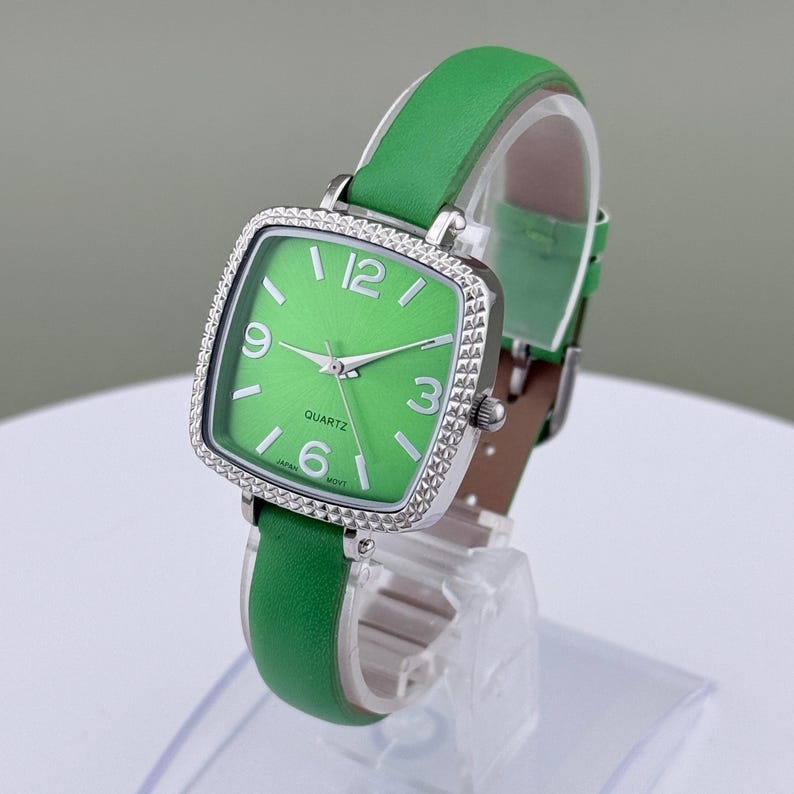 May include: A green quartz watch with a square face and a silver bezel. The watch has a green leather strap and a white dial with black numerals. The watch is displayed on a clear stand.