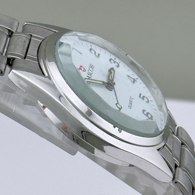 Pu&ograve; includere: Un orologio da polso argentato con quadrante bianco e numeri neri. L'orologio ha un movimento al quarzo e un cinturino in metallo. Il quadrante mostra il marchio "ANCCHI". L'orologio &egrave; angolato, mostrando il quadrante e il cinturino.
