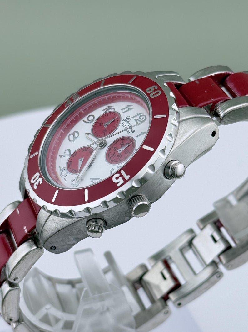May include: A silver-toned wristwatch with a red bezel and accents. The watch face is white with red sub-dials and hands. The watch band is a combination of silver and red links. The watch has a chronograph function.