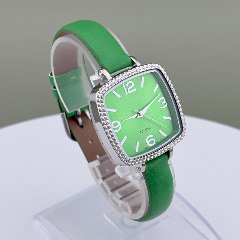 May include: A green quartz watch with a square face and silver bezel. The watch has a green leather strap and a light green watch face with white numerals. The watch is displayed on a clear stand.