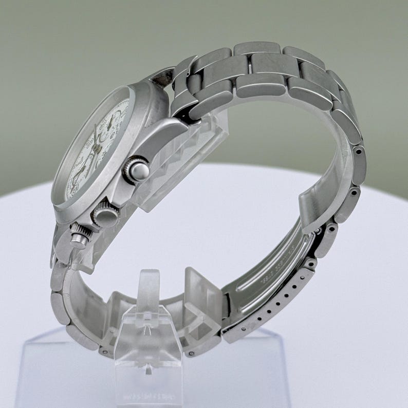 Puede incluir: Un reloj de pulsera plateado con esfera blanca y correa de metal eslabonada. El reloj tiene una esfera redonda con agujas e indicadores visibles. La correa es curva y el reloj se exhibe sobre un soporte transparente.