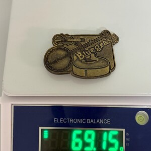 May include: A bronze-colored pin featuring a banjo and a violin, with the word "Bluegrass" inscribed. The pin is on a scale, weighing 69.15 grams. The scale is white and has a digital display.