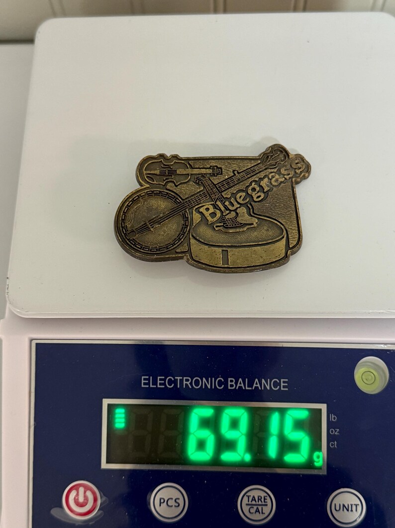 May include: A bronze-colored pin featuring a banjo and a violin, with the word "Bluegrass" inscribed. The pin is on a scale, weighing 69.15 grams. The scale is white and has a digital display.