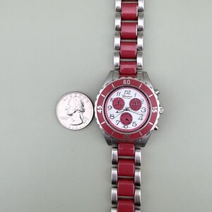 May include: A Geneva Platinum wristwatch with a red and silver band and a white face. The watch has a red bezel and three small dials. A quarter is next to the watch for size comparison.