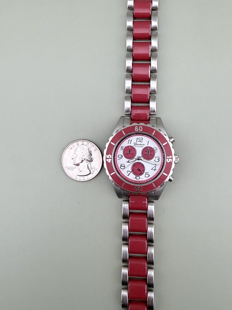 May include: A Geneva Platinum wristwatch with a red and silver band and a white face. The watch has a red bezel and three small dials. A quarter is next to the watch for size comparison.