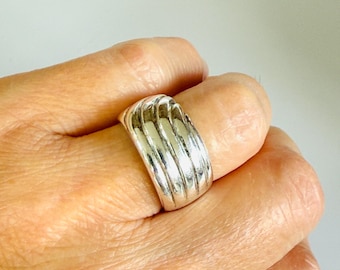 May include: A silver-colored ring with a ribbed design. The ring is wide and has a series of vertical ridges across the top. The ring is worn on a finger.