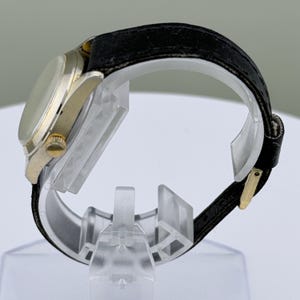 K&ouml;nnte beinhalten: Seitenansicht einer goldfarbenen Armbanduhr mit schwarzem Lederarmband. Die Uhr hat ein rundes Zifferblatt und eine goldene Krone. Das Armband wird mit einer goldfarbenen Schnalle befestigt. Die Uhr wird auf einem transparenten Kunststoffst&auml;nder ausgestellt.