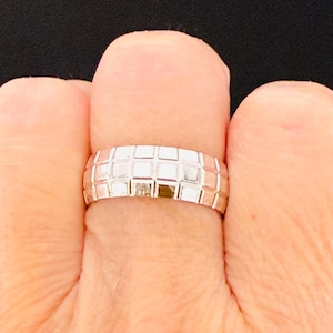May include: A silver ring with a textured, grid-like pattern. The ring is shown on a finger, with a close-up view highlighting the geometric design. The ring's surface reflects light, creating a bright, shiny appearance. The ring is a piece of jewelry.