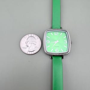 May include: A square-faced quartz watch with a light green dial and a matching green leather strap. The watch has silver-coloured trim and a white dial with the numbers 3, 6, 9, and 12. A U.S. quarter is next to the watch.
