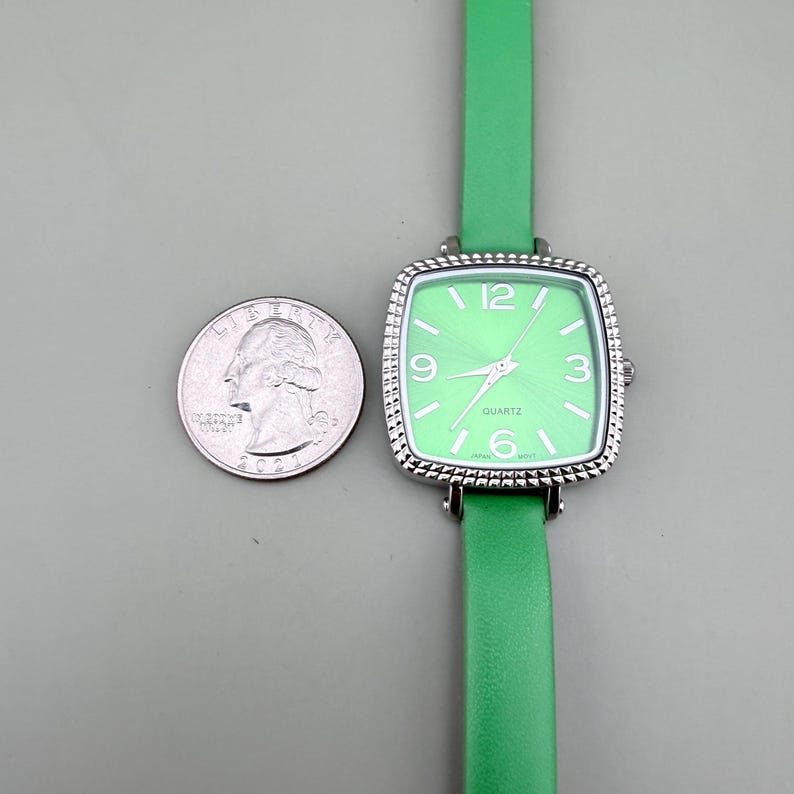 May include: A square-faced quartz watch with a light green dial and a matching green leather strap. The watch has silver-coloured trim and a white dial with the numbers 3, 6, 9, and 12. A U.S. quarter is next to the watch.