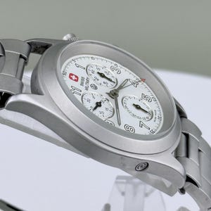 Puede incluir: Un reloj Swiss Military plateado con esfera blanca. El reloj presenta m&uacute;ltiples subesferas, una pantalla de fecha y el logotipo Swiss Military. Tiene una correa de acero inoxidable y una caja redonda.
