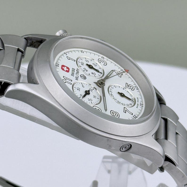 Puede incluir: Un reloj Swiss Military plateado con esfera blanca. El reloj presenta m&uacute;ltiples subesferas, una pantalla de fecha y el logotipo Swiss Military. Tiene una correa de acero inoxidable y una caja redonda.