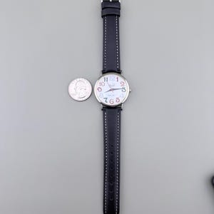 May include: A wristwatch with a black leather band and a silver buckle. The watch face is white with colorful numbers and hands. A U.S. quarter is next to the watch for size comparison.