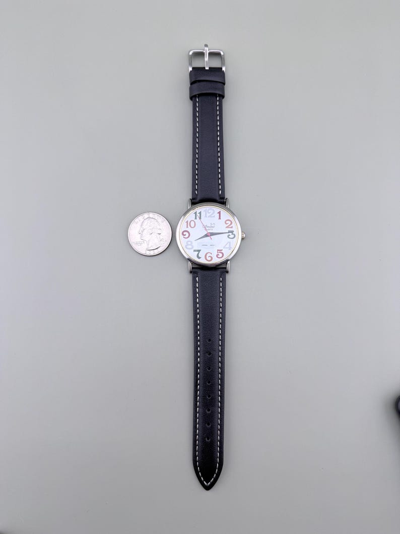 May include: A wristwatch with a black leather band and a silver buckle. The watch face is white with colorful numbers and hands. A U.S. quarter is next to the watch for size comparison.