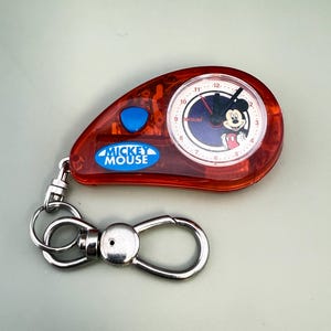 May include: Red Mickey Mouse keychain watch with a silver clasp. The watch face features Mickey Mouse and the text "Mickey Mouse". The watch has a blue button and a silver key ring.