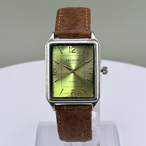 May include: A vintage Helbros quartz watch with a rectangular silver-tone case and a brown leather strap. The watch face is a light green color with black numerals and hands. The watch face has the text "HELBROS Since 1913" and "QUARTZ".