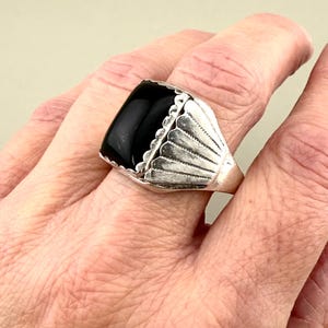 May include: A silver ring with a large, rectangular black stone. The ring has a decorative, fan-like design around the stone. The ring is on a person's finger.