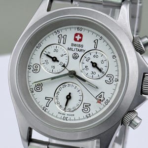 Puede incluir: Un reloj Swiss Military plateado con esfera blanca. El reloj presenta n&uacute;meros negros, tres subesferas y un logotipo de cruz suiza roja. Tiene una correa de acero inoxidable y funci&oacute;n cron&oacute;grafo.